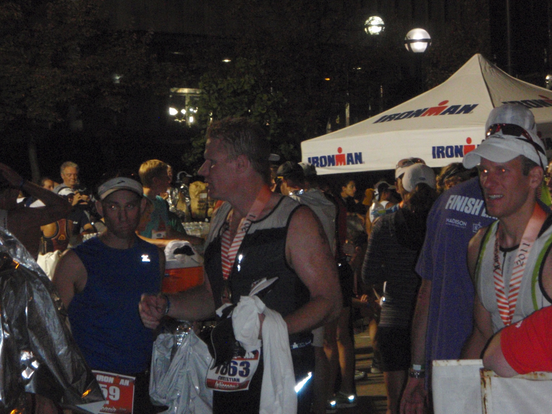 Ironman Wisconsin finish chute at night; athletes with medals and space blankets in the crowd.