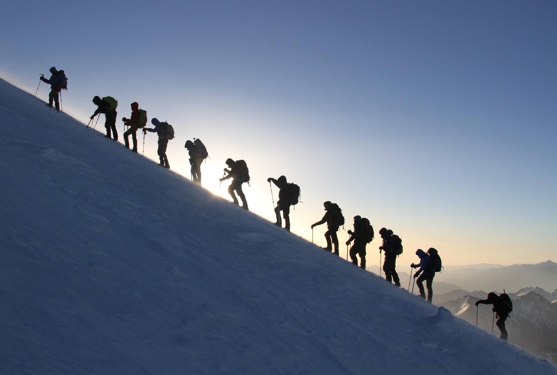 Rope team climbing single file on Elbrus at dawn, snow slope with alpenglow on the summit ridge.